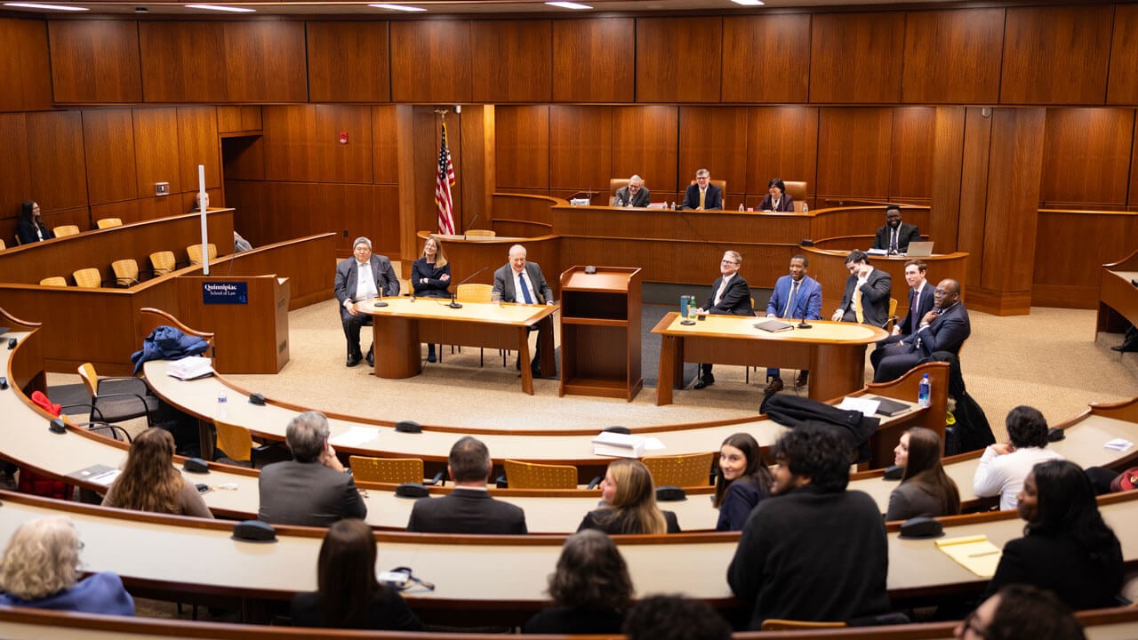 A panel at the front of a courtroom speaking among themselves as the audience watches