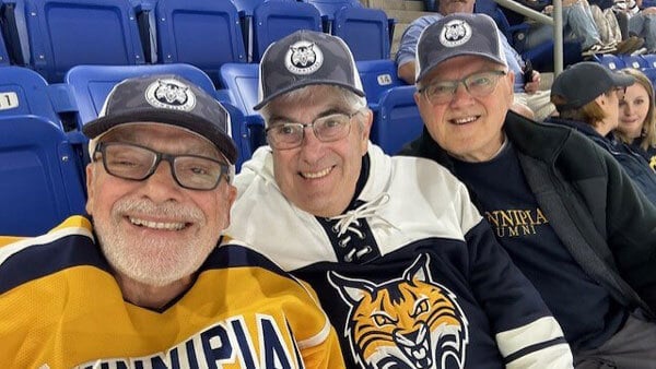 group of people smiling wearing matching Quinnipiac hockey hat sitting in blue stadium seats