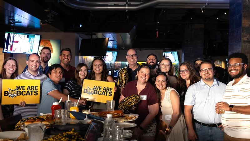 A dozen alumni and faculty hold Bobcats signs and smile for a photo in a bar