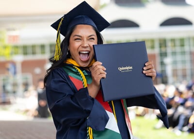 A graduate in her cap and gown excitedly holds up her diploma cover during Commencement