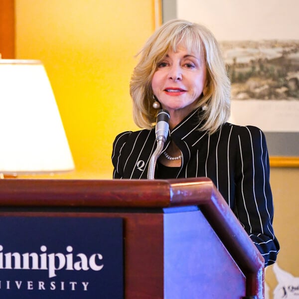 President Judy Olian speaks at a podium with a Quinnipiac University logo