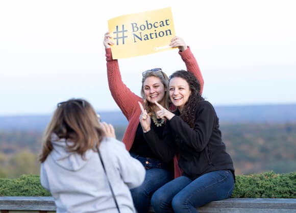 Two alumni pose for a photo sitting on a wall at Rocky Top holding a #Bobcat Nation sign