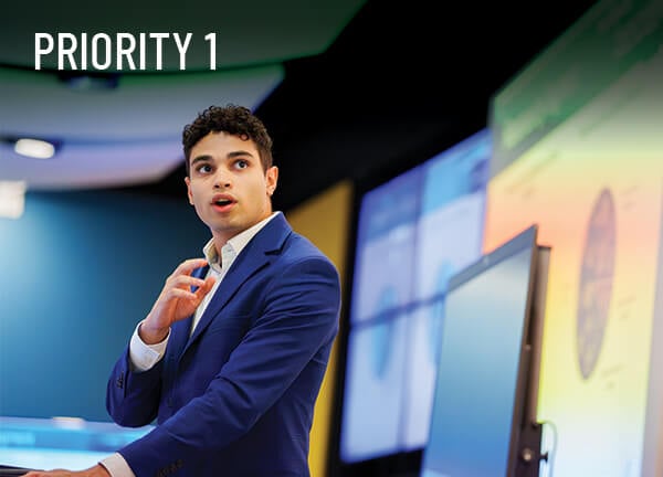 Priority 1: A student in a suit stands a podium and points to his presentation on screen