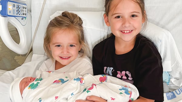 Two little girls hold their new-born brother on a hospital bed