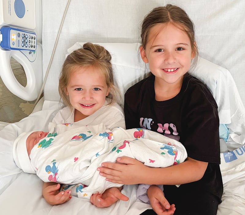 Two little girls hold their new-born brother on a hospital bed