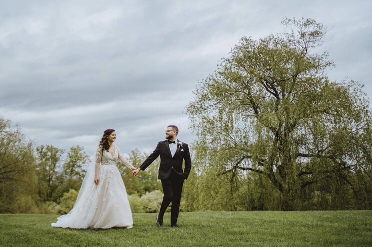 Andy Thane and Amy (Brenner) Thane hold hands and walk in front of a row of trees on their wedding day
